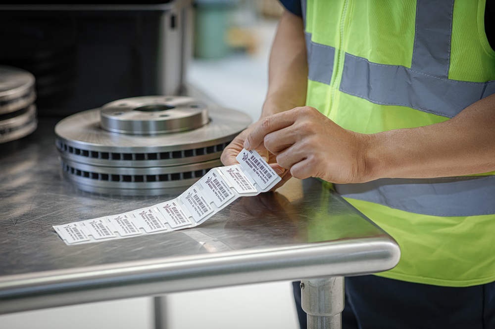 En person i arbeidsklær holder utskrevne etiketter ved et metallbord med maskindeler i bakgrunnen.