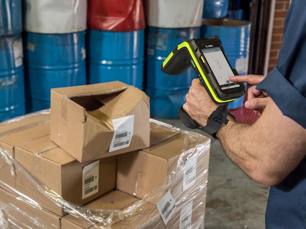 En mann i et lager skanner en pakke ved hjelp av en Zebra RFD90 RFID-enhet. Skjermen på enheten viser et applikasjonsgrensesnitt, og han berører skjermen med høyre hånd. Bakgrunnen inkluderer flere stablede esker og blå industrielle tønner.